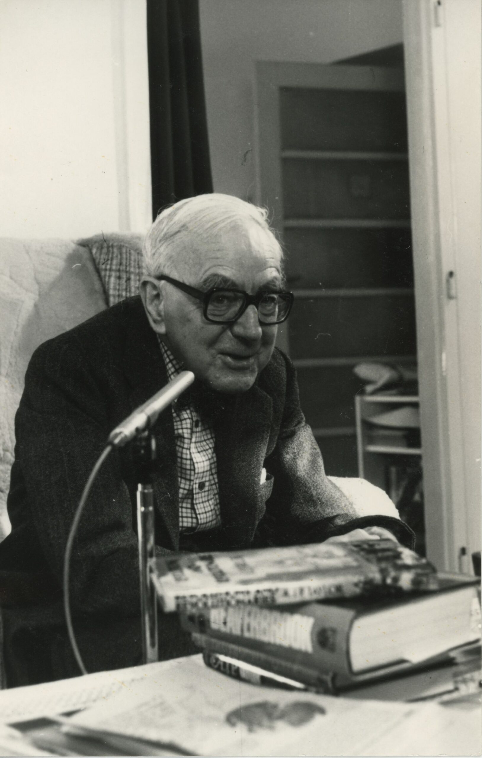 A.J.P. Taylor sitting at a desk, with a microphone positioned by his face. He wears thick-rimmed spectacles, a suit a tie, with his trademark bow tie.